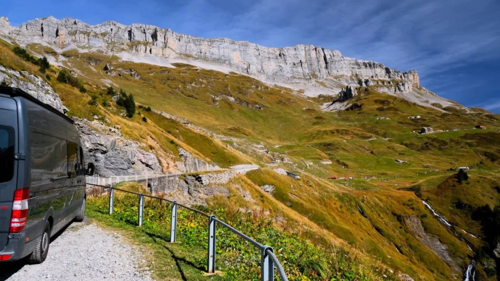 Roadtrip Europa: Schwarzer Camper Van steht auf Schotterparkplatz in den Schweizer Alpen, Blick auf gewaltige Felswand und grünes Tal am Klausenpass